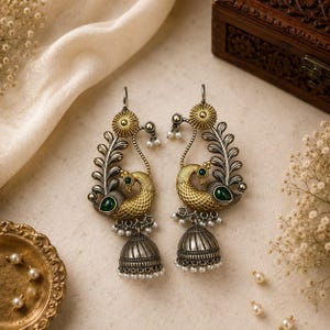 May include: A pair of oxidized silver and gold-toned peacock-shaped earrings featuring green stone accents, intricate textured metalwork, and dangling bell-shaped jhumka charms adorned with small white beads along the bottom edges.