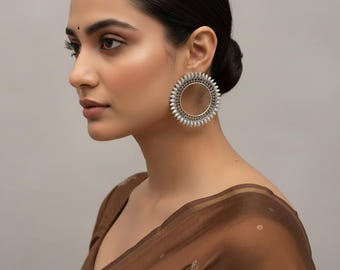 May include: Silver-toned hoop earrings with a sunburst design. The earrings feature a circular frame with small, pointed embellishments around the outer edge. The earrings are worn by a person with dark hair and a brown top.