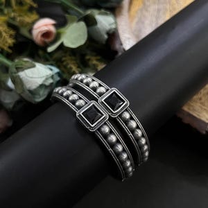 May include: A set of three silver bangles with a dark finish. Each bangle features a row of small, round beads and a square, black gemstone set in a square frame. The bangles are displayed on a black cylindrical surface.
