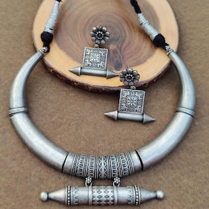 Puede incluir: Un collar de plata con un colgante de media luna y pendientes a juego. El collar tiene una banda decorativa con diseños intrincados. Los pendientes tienen un colgante cuadrado con un diseño floral.