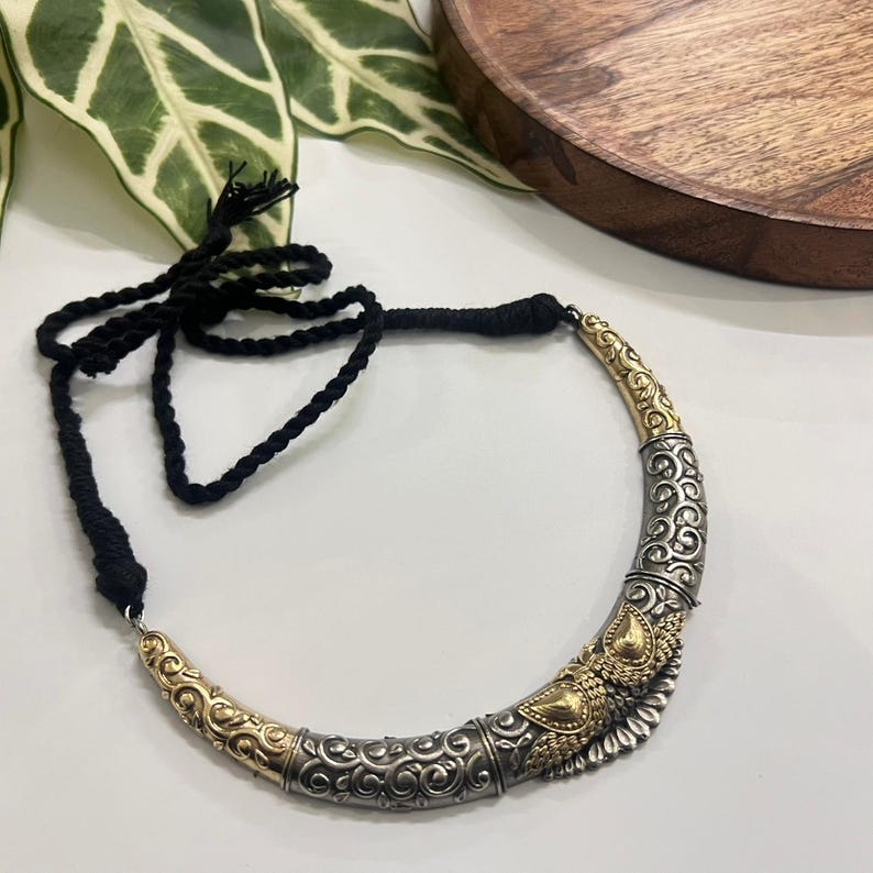 Puede incluir: Un collar plateado y dorado con un dise&ntilde;o curvo y ornamentado. El collar presenta patrones intrincados y un cord&oacute;n de cuerda negro. El trabajo en metal tiene una est&eacute;tica vintage, con acabados pulidos y oxidados. El collar es una pieza llamativa.