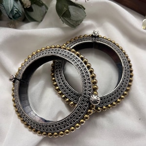 Könnte beinhalten: Zwei silber- und goldfarbene Armreifen mit aufwendigen Details. Die Armreifen haben eine silberne Basis mit einem dekorativen Muster und sind mit goldfarbenen Perlen an den Rändern verziert. Der Schmuck wird auf weißem Stoff präsentiert.