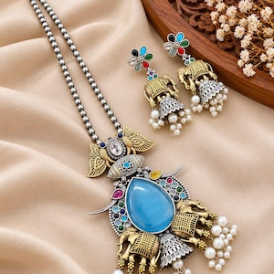 May include: A silver-toned beaded necklace with a large pendant featuring a central blue teardrop stone, gold-toned elephant motifs, and dangling white pearls. Matching drop earrings feature gold-toned elephant figures, colorful floral accents, and white pearl beads.
