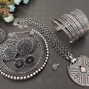 Può includere: Un set di gioielli in argento con una collana, orecchini, un braccialetto e un ciondolo. La collana ha un grande ciondolo rotondo con disegni intricati. Anche gli orecchini sono rotondi con disegni intricati. Il braccialetto è un braccialetto spesso con più strati. Il ciondolo è un grande ciondolo rotondo in filigrana.