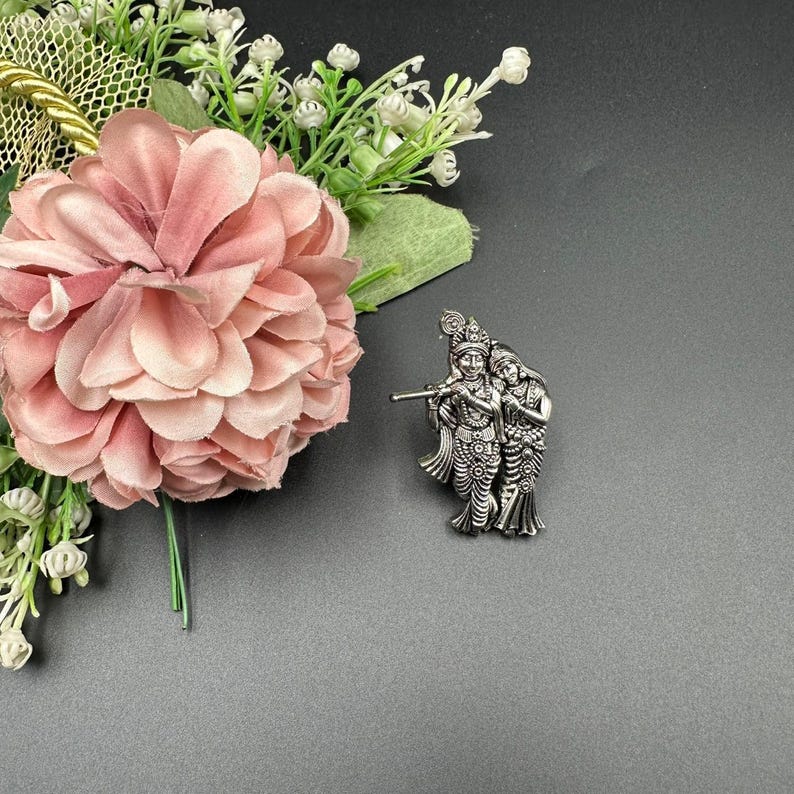 Puede incluir: Broche de metal plateado que representa dos figuras, una tocando la flauta. El broche est&aacute; sobre un fondo gris, con una flor rosa y detalles florales blancos en la esquina superior izquierda.