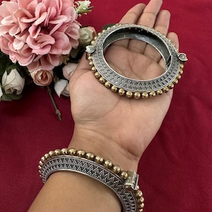 May include: Two silver and gold bangles with detailed patterns and beaded embellishments. The bangles are shown on a red surface, with pink flowers in the background. The jewellery has a vintage or ethnic style.