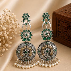 May include: A pair of silver-toned dangle earrings featuring green circular stones, intricate metalwork with gold-toned bird motifs, and a row of small white beads hanging from the bottom of each large circular pendant.