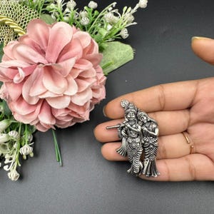 Puede incluir: Un colgante plateado con dos figuras, una tocando una flauta. Las figuras est&aacute;n adornadas con ropa y tocados detallados. El colgante se sostiene en una mano, con una flor rosa y vegetaci&oacute;n en el fondo.