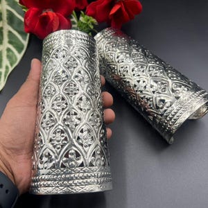 Könnte beinhalten: Zwei silberfarbene, zylindrische Kerzenhalter mit aufwendigen, durchbrochenen Mustern. Einer steht aufrecht, der andere liegt auf der Seite. Die Halter haben ein detailliertes Blumendesign und sind wahrscheinlich aus Metall. Der Hintergrund umfasst rote Blumen.