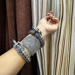 以下が含まれることがあります： シルバーカラーのブレスレットとカフスのコレクション。ブレスレットは、中央にサンバーストのデザインが施された幅広のカフスと、小さくテクスチャ加工されたバンドなど、複雑なパターンが特徴です。茶色とベージュのストライプのカーテンを背景に、手首にジュエリーが飾られています。