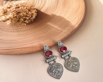 May include: A pair of silver-toned earrings with a vintage design. Each earring features a deep red oval stone, a decorative cylindrical element, and a heart-shaped pendant with intricate patterns. The earrings rest on a light wooden surface.