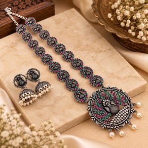 May include: A silver-toned necklace and earring set featuring oxidized metal with embedded green and pink stones. The necklace has a circular peacock pendant with dangling white beads, and the matching jhumka earrings also feature white bead accents.