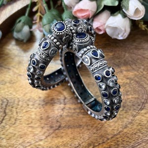 Può includere: Due braccialetti d'argento con dettagli intricati e pietre preziose blu e trasparenti incastonate. I braccialetti sono esposti su una superficie di legno, con una composizione floreale sullo sfondo. I gioielli hanno un'estetica antica o vintage.