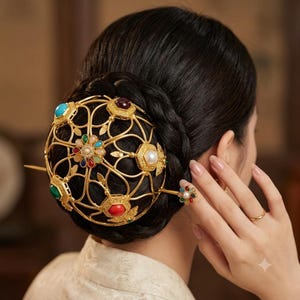 Pode incluir: Um acessório de cabelo em metal dourado com um design de gaiola esférica vazada, adornado com várias pedras cabochão coloridas, incluindo turquesa, vermelho e verde, juntamente com uma pérola central e um grampo de cabelo decorativo.