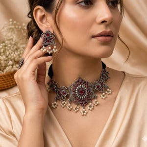 Puede incluir: Un conjunto de joyas plateadas con collar, pendientes y anillo. El collar y los pendientes tienen un diseño floral con detalles de piedras preciosas rojas y perlas de imitación colgantes. El anillo también tiene un diseño similar.