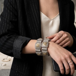 Könnte beinhalten: Silber- und goldfarbene Metallarmbänder mit quadratischem Design und floralen Akzenten. Die Person trägt einen schwarzen Blazer und hält ein schwarzes Smartphone. Ein silberner Ring befindet sich am Finger.