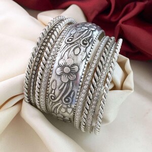 May include: A collection of silver bangles with detailed floral and leaf patterns. The central bangle showcases a prominent flower, complemented by twisted rope and beaded designs on the other bangles. They are presented on a cream fabric.