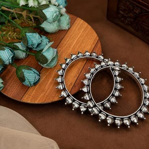 Può includere: Due braccialetti color argento con un design a punte sono esposti su una superficie marrone. I braccialetti sono accanto a un piatto di legno con fiori blu e verdi. Una scatola di legno è sullo sfondo.