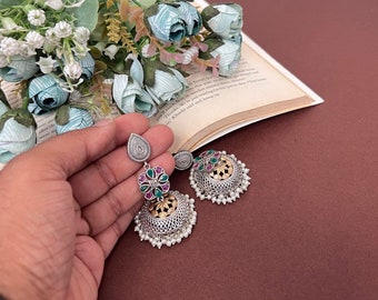 May include: Silver-toned earrings with a teardrop top and a floral design with red and green accents. The earrings have a bell-shaped bottom with gold detailing and small white beads. The earrings are held in a hand.