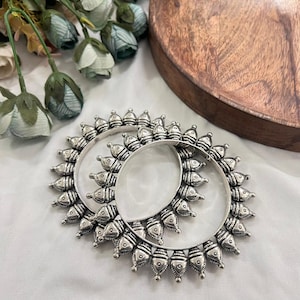 May include: A pair of silver bangles with a detailed, sunburst design. Each bangle has small, decorative elements around the circumference. The jewellery is displayed on a white surface, with floral accents in the background.