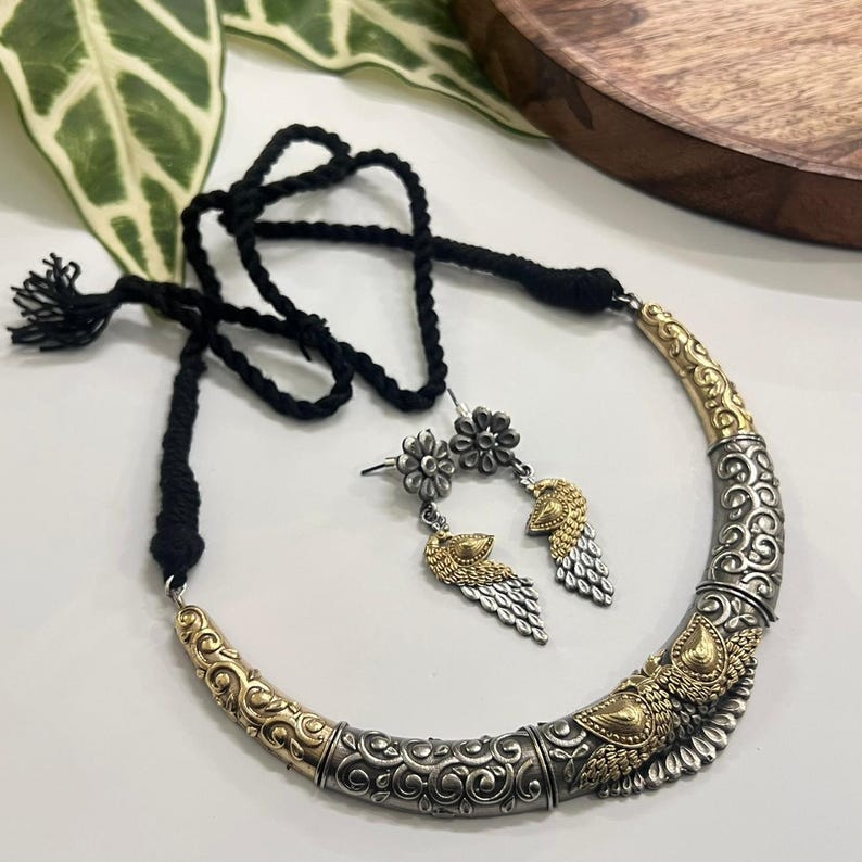 Puede incluir: Un conjunto de collar y pendientes plateados y dorados. El collar tiene un dise&ntilde;o curvo con patrones intrincados y un cord&oacute;n negro. Los pendientes presentan un perno floral y un colgante en forma de ala. Las joyas se exhiben sobre una superficie blanca.