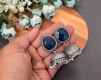 May include: A pair of silver-toned jhumka earrings featuring a faceted, dark blue oval stone at the top and a bell-shaped base adorned with intricate metal engravings and small white beads hanging from the bottom edge.