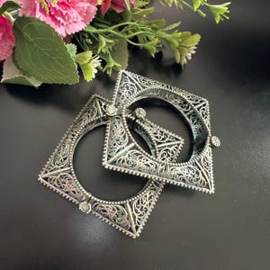 May include: Two silver square bangles with detailed filigree patterns. The bangles have a hinged clasp. The background is black with pink and green floral accents. Each bangle has an open centre.