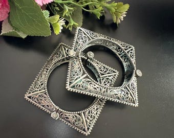May include: Two silver-toned square bangles with intricate filigree designs. The bangles are open in the center and have a hinged clasp. The background is black with pink and green floral accents.