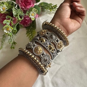 May include: A collection of silver and gold-coloured bangles are worn on a wrist. The bangles showcase detailed designs, including circular patterns, embossed features, and elephant motifs. The jewellery is set against a soft, white backdrop, with a bouquet of pink and white flowers in the background.