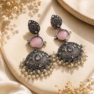 May include: A pair of oxidized silver-tone dangle earrings featuring a teardrop-shaped top, a round faceted pink stone in the center, and a large dome-shaped base with intricate floral engravings, clear crystals, and small white pearl-like beads.