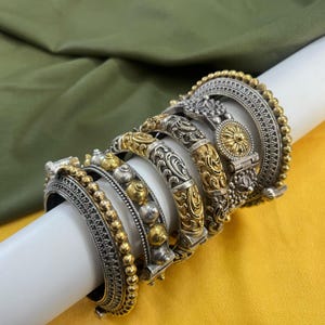 Op de afbeelding: Een set van zes zilveren en gouden armbanden met ingewikkelde ontwerpen. De armbanden zijn op elkaar gestapeld en hebben verschillende texturen en patronen.