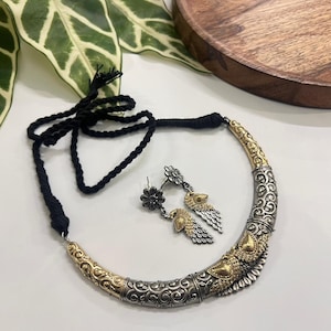 Puede incluir: Un conjunto de collar y pendientes plateados y dorados. El collar tiene un cord&oacute;n negro y un colgante curvo y adornado. Los pendientes presentan dise&ntilde;os florales y en forma de plumas. Las joyas se exhiben sobre una superficie blanca.