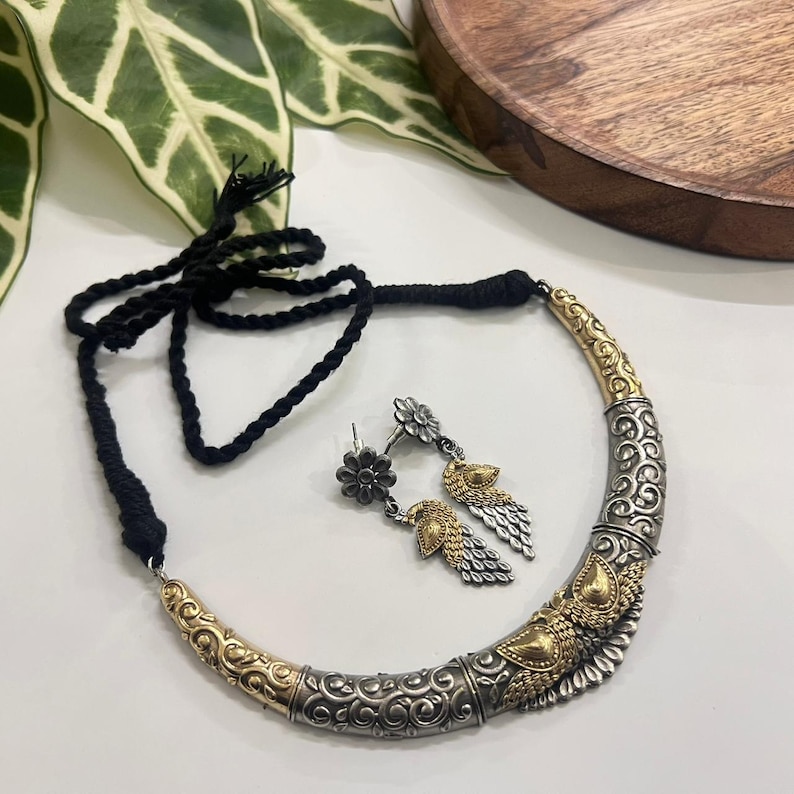 Puede incluir: Un conjunto de collar y pendientes plateados y dorados. El collar tiene un cord&oacute;n negro y un colgante curvo y adornado. Los pendientes presentan dise&ntilde;os florales y en forma de plumas. Las joyas se exhiben sobre una superficie blanca.