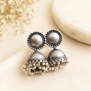 May include: A pair of oxidized silver jhumka earrings featuring a circular stud top with a beaded border and a bell-shaped drop base adorned with numerous small white seed beads hanging from the bottom edge of the metal dome structure.