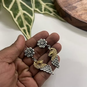 Puede incluir: Pendientes de pavo real plateados y dorados. Cada pendiente presenta un perno de flor plateado, una cabeza de pavo real dorada y una cola plateada. Los pendientes se sostienen en una mano sobre un fondo neutro. Joyer&iacute;a &uacute;nica.
