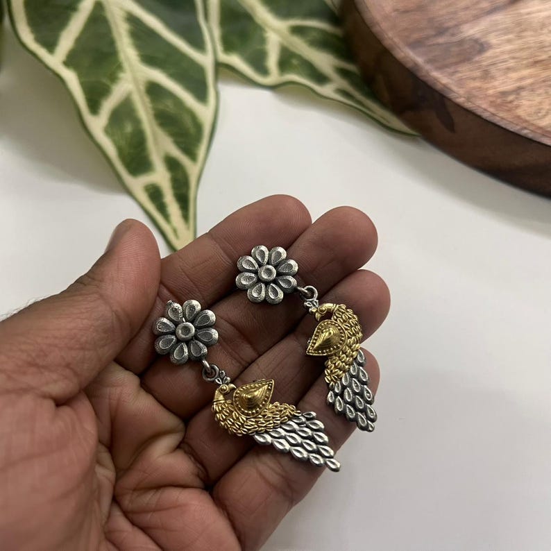 Puede incluir: Pendientes de pavo real plateados y dorados. Cada pendiente presenta un perno de flor plateado, una cabeza de pavo real dorada y una cola plateada. Los pendientes se sostienen en una mano sobre un fondo neutro. Joyer&iacute;a &uacute;nica.