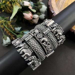 Può includere: Una collezione di braccialetti argentati con intricati disegni di elefanti e intrecci. I braccialetti sono esposti su una forma cilindrica nera, mostrando la loro dettagliata lavorazione artigianale. I motivi degli elefanti aggiungono un tocco culturale ai gioielli.