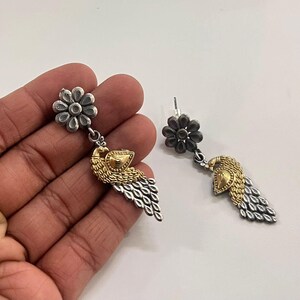 Puede incluir: Pendientes colgantes plateados y dorados. Cada pendiente presenta un perno en forma de flor, un dise&ntilde;o de p&aacute;jaro dorado y una gota plateada en forma de pluma. Los pendientes se muestran sobre un fondo blanco.