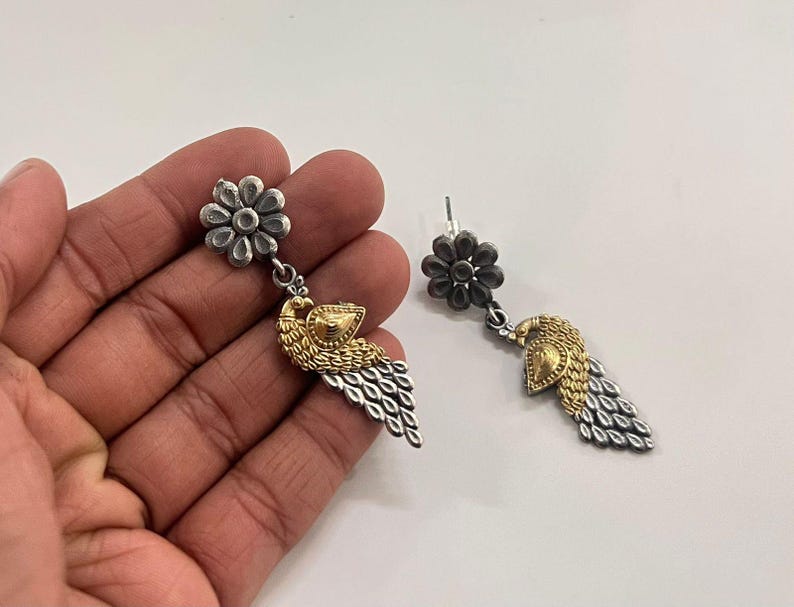 Puede incluir: Pendientes colgantes plateados y dorados. Cada pendiente presenta un perno en forma de flor, un dise&ntilde;o de p&aacute;jaro dorado y una gota plateada en forma de pluma. Los pendientes se muestran sobre un fondo blanco.