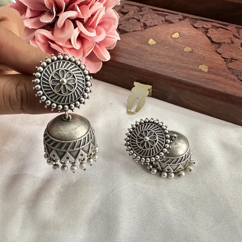 May include: Silver-toned jhumka earrings with a floral design and small dangling beads. The earrings have a bell shape with geometric patterns and are displayed on a white surface.