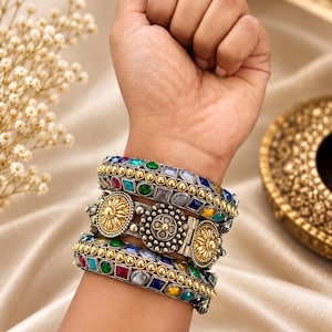 Könnte beinhalten: Ein Set aus drei gestapelten Metallarmreifen in Gold- und Silbertönen. Die Armbänder sind mit bunten quadratischen und ovalen Steinen in Rot, Blau, Grün und Gelb sowie mit detaillierten floralen Metallverzierungen versehen.