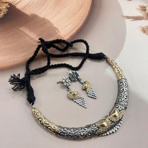 Puede incluir: Un conjunto de collar y pendientes plateados y dorados. El collar tiene un cord&oacute;n negro y un colgante curvo y adornado. Los pendientes presentan un perno floral y una gota en forma de pluma. Las joyas se exhiben sobre una superficie neutra.