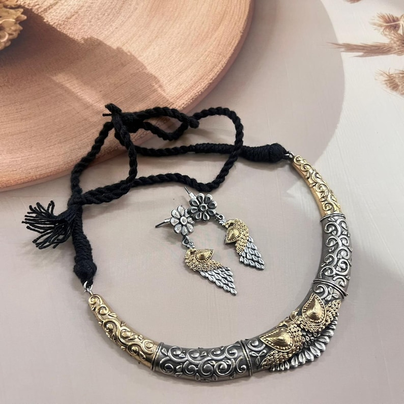 Puede incluir: Un conjunto de collar y pendientes plateados y dorados. El collar tiene un cord&oacute;n negro y un colgante curvo y adornado. Los pendientes presentan un perno floral y una gota en forma de pluma. Las joyas se exhiben sobre una superficie neutra.