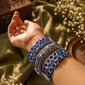 Könnte beinhalten: Silberfarbene Armbänder mit blauen Edelsteinakzenten. Die Armbänder weisen aufwendige Designs auf, darunter florale und geometrische Muster. Die Edelsteine sind tiefblau und die Armbänder werden am Handgelenk präsentiert.