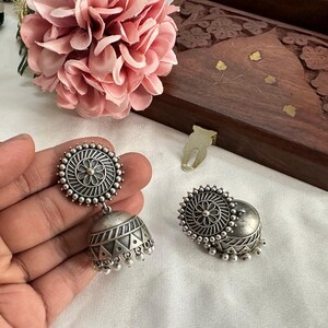 May include: A pair of silver-toned jhumka earrings featuring a circular stud with a central flower motif and radiating lines, connected to a bell-shaped drop decorated with geometric triangle patterns and small dangling beads.