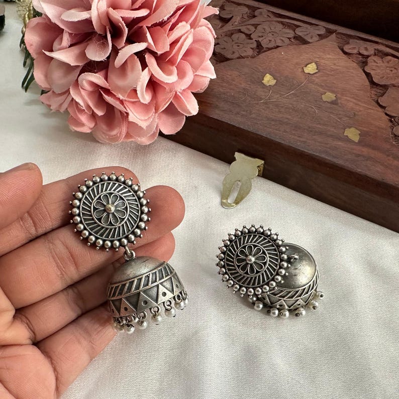 May include: A pair of silver-toned jhumka earrings featuring a circular stud with a central flower motif and radiating lines, connected to a bell-shaped drop decorated with geometric triangle patterns and small dangling beads.