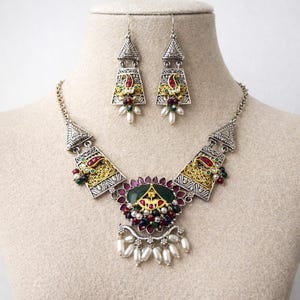 May include: A silver-toned necklace and earring set featuring intricate metalwork with gold-tone accents. The jewellery includes dark green, red, and blue beads, along with white teardrop-shaped dangles. The central pendant displays a large green stone.