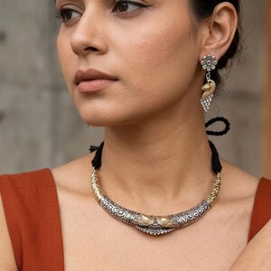 Puede incluir: Un collar plateado y dorado con un dise&ntilde;o ornamentado, combinado con pendientes a juego. Los pendientes presentan un perno en forma de flor y un dise&ntilde;o colgante inspirado en un pavo real. La joyer&iacute;a la lleva una persona que lleva una camiseta de color &oacute;xido.