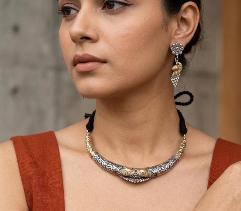 Puede incluir: Un collar plateado y dorado con un dise&ntilde;o ornamentado, combinado con pendientes a juego. Los pendientes presentan un perno en forma de flor y un dise&ntilde;o colgante inspirado en un pavo real. La joyer&iacute;a la lleva una persona que lleva una camiseta de color &oacute;xido.