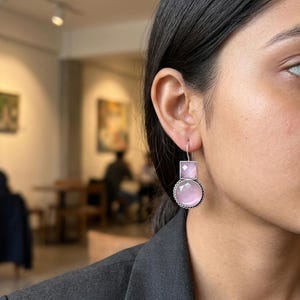 May include: Silver earrings featuring a square and a round, pale pink gemstone. The earrings hang down and have a decorative border around the round stone. The earrings are worn on a person's ear.